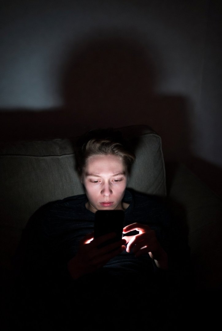 A person lying on a couch scrolling their phone in the dark, face lit by screen glow—eyes tired but still scrolling.