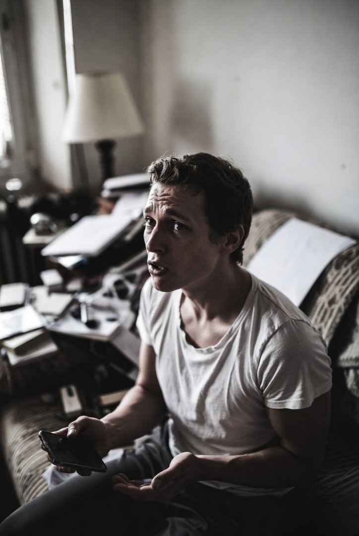 A person sitting on a couch, looking slightly overwhelmed, phone in hand, messy room around them