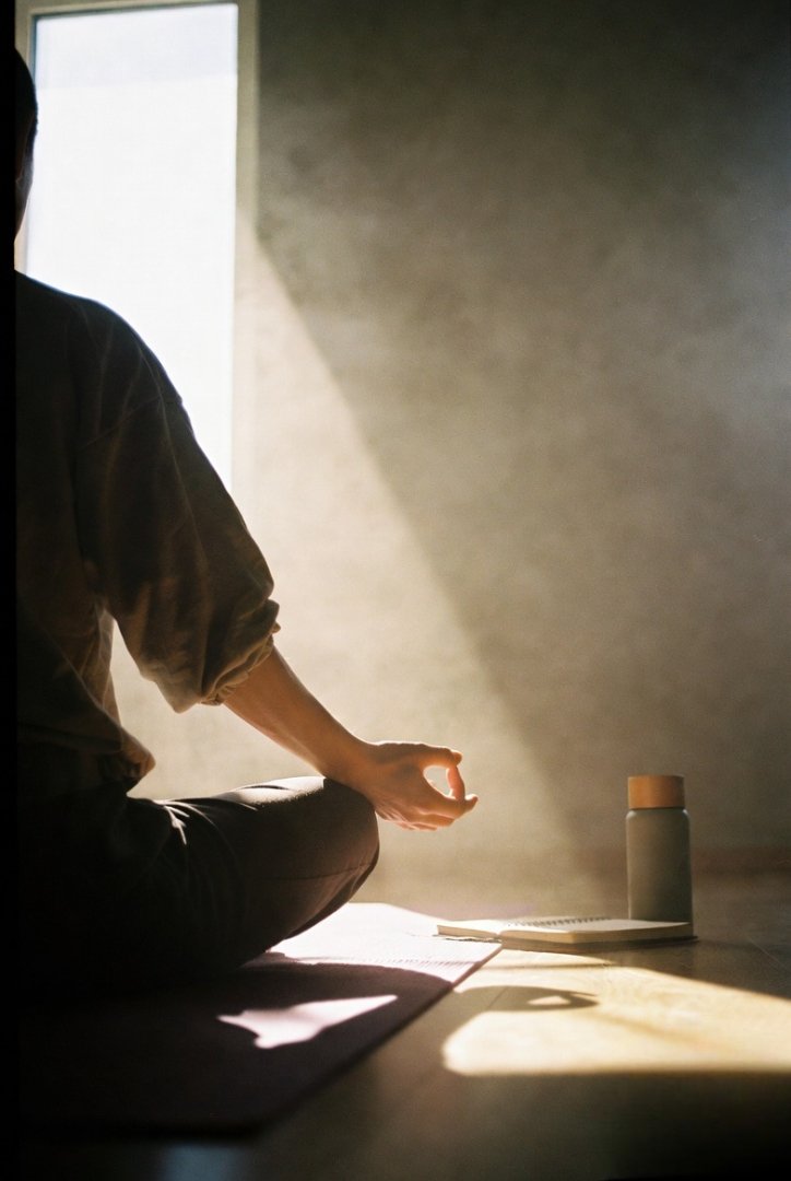 a person sitting cross-legged on a yoga mat near a window, sunlight spilling in