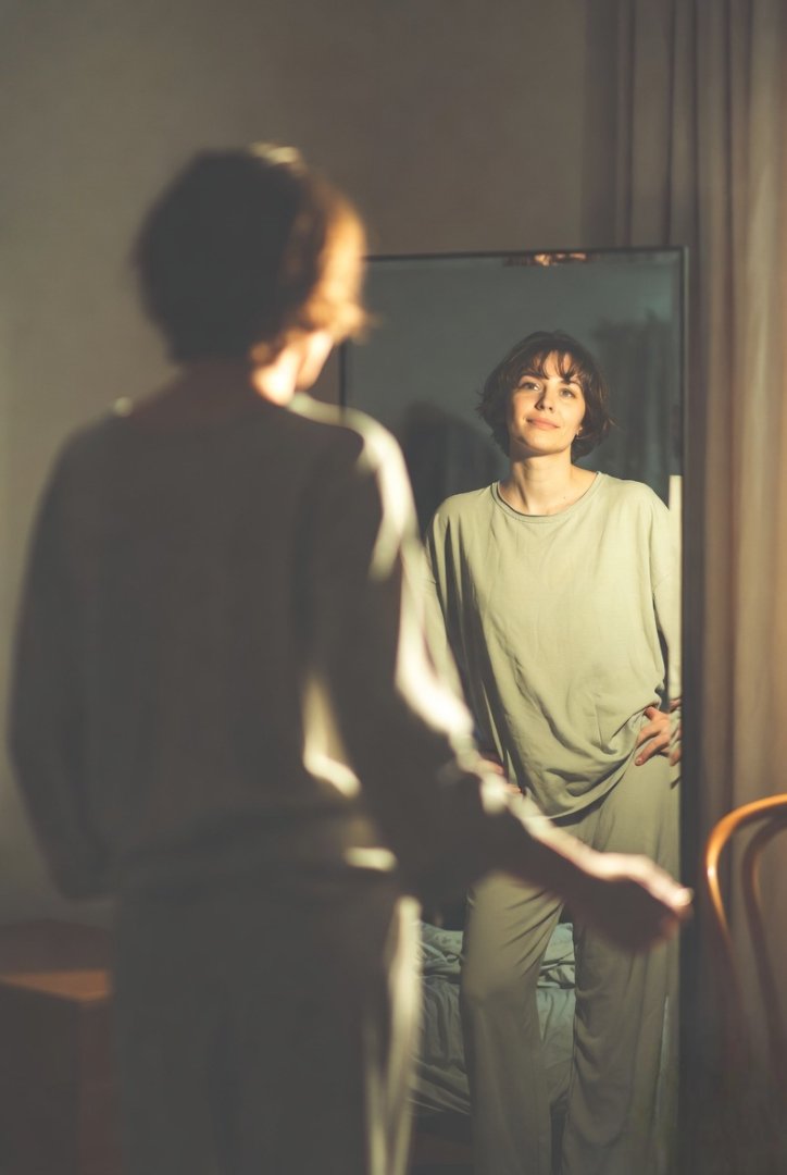 A person standing in front of a mirror, soft morning light, slightly messy room behind them but a calm